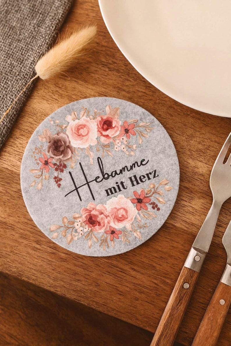 May include: A circular light gray felt coaster featuring a floral wreath design in shades of pink, peach, and brown. The text "Hebamme mit Herz" is printed in black script and serif font in the center of the floral arrangement on the coaster.