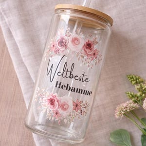 May include: A clear glass tumbler with a bamboo lid and a clear glass straw. The glass features a floral design in shades of pink and green, with the black text "Weltbeste Hebamme" printed in the center between the floral arrangements.