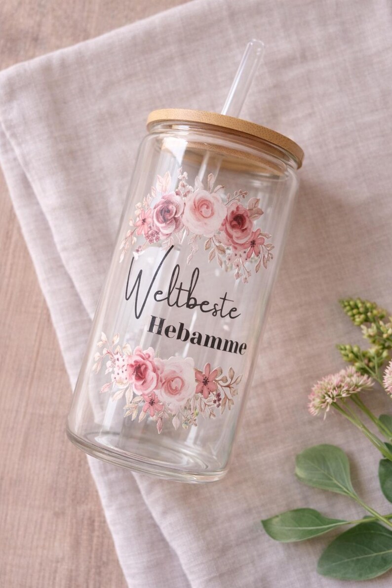 May include: A clear glass tumbler with a bamboo lid and a clear glass straw. The glass features a floral design in shades of pink and green, with the black text "Weltbeste Hebamme" printed in the center between the floral arrangements.