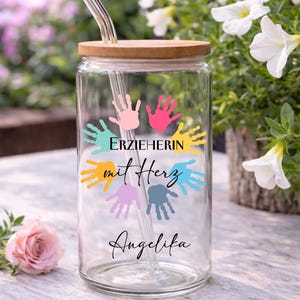May include: A clear glass tumbler with a bamboo lid and a glass straw. The tumbler is decorated with colorful handprints and the text "ERZIEHERIN mit Herz Angelika".