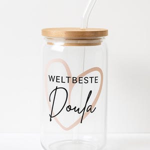 May include: A clear glass tumbler with a bamboo lid and a glass straw. The front features a light pink heart outline with the black text WELTBESTE and the word Doula written in a cursive script font centered within the heart shape.