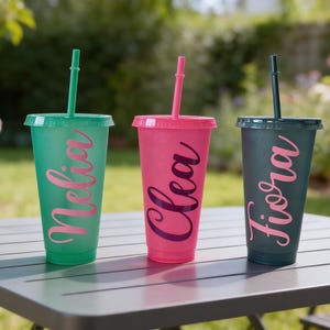 Puede incluir: Tres vasos de pl&aacute;stico reutilizables con tapas y pajitas en verde, rosa y gris oscuro. Cada vaso tiene un acabado brillante y un nombre personalizado en letra cursiva: Nelia en el vaso verde, Clea en el rosa y Fiora en el vaso de color gris oscuro.