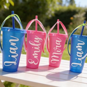 Puede incluir: Cuatro vasos de pl&aacute;stico con purpurina, tapas y pajitas, cada uno con un nombre personalizado en letras blancas. Dos vasos azules llevan el nombre Liam, mientras que dos vasos rosas llevan los nombres Emily y Mira. Pajitas decoradas con cintas.