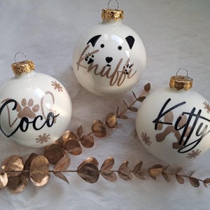 May include: Three white Christmas baubles with gold caps. Each bauble has a pet's name in black script with a paw print and snowflakes. One bauble has a dog face design. The names are "Coco", "Knuff", and "Kitty".