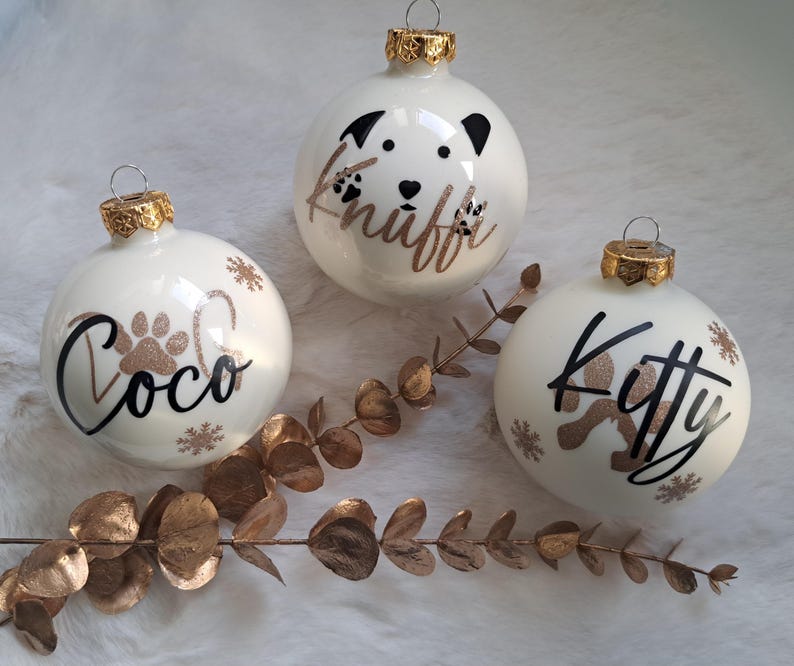 May include: Three white Christmas baubles with gold caps. Each bauble has a pet's name in black script with a paw print and snowflakes. One bauble has a dog face design. The names are "Coco", "Knuff", and "Kitty".