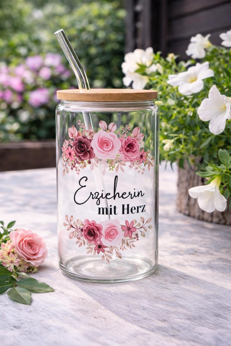 Op de afbeelding: Een doorzichtig glazen beker met een bamboe deksel en een glazen rietje, voorzien van een bloemmotief met roze en bordeauxrode rozen en bladeren. De tekst "Erzieherin mit Herz" staat in zwarte schrijf- en schreefletters in het midden.