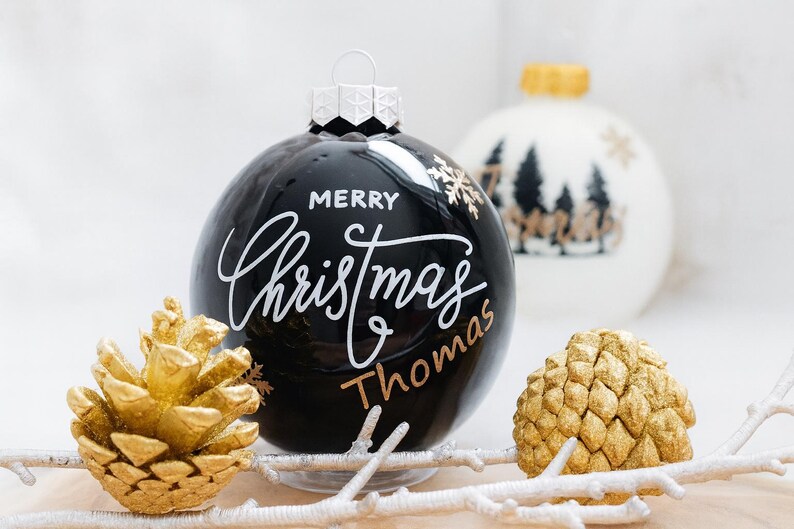 以下が含まれることがあります： 「Merry Christmas Thomas」と書かれた白い文字が入った黒いクリスマスオーナメント。金の雪の結晶と金色の松ぼっくりも見える。背景には森の風景が描かれた白いオーナメントがある。