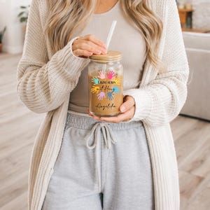 May include: A clear glass tumbler with a wooden lid and a straw, filled with a light brown beverage. The tumbler is decorated with colorful handprints and text that reads "Erzieherin mit Herz Angelina."