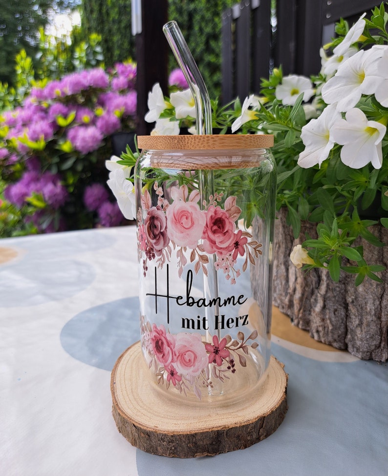 May include: A clear glass tumbler with a bamboo lid and a bent glass straw. The glass features a floral design in shades of pink and burgundy, with the black text "Hebamme mit Herz" printed in the center between the floral arrangements.