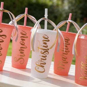 Puede incluir: Cinco vasos de pl&aacute;stico reutilizables con tapas y pajitas, cuatro en coral y uno en blanco, con acabado brillante y letras doradas en cursiva que dicen Franzi, Simone, Christine, Martina y Katharina, decorados con cintas atadas.
