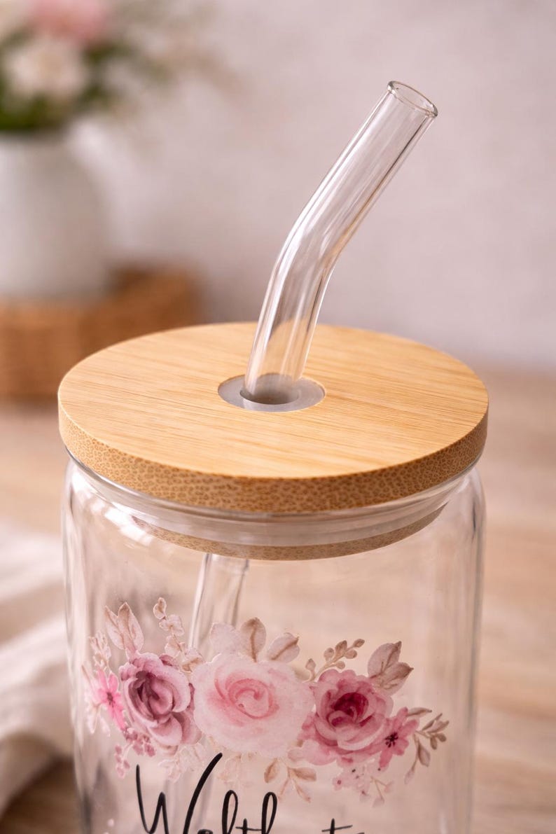 May include: A clear glass tumbler featuring a floral design with pink and mauve roses, topped with a circular bamboo lid and a bent clear glass straw. The floral pattern is printed on the front of the glass, which has a smooth, cylindrical shape.