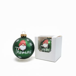 K&ouml;nnte beinhalten: Eine gr&uuml;ne Weihnachtskugel mit einem Zwergen-Design und dem Namen "Thomas" in wei&szlig;er Schrift. Der Schmuck hat eine goldene Kappe und steht auf einem transparenten St&auml;nder. Eine zweite Kugel mit dem Namen "Sophie" ist in einer wei&szlig;en Box zu sehen.