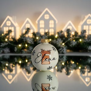 以下が含まれることがあります： 白いクリスマスオーナメント。名前「Elena」が筆記体で書かれ、鹿のイラストが描かれています。オーナメントは金色の金属製のキャップが付いており、黒い雪の結晶で飾られています。背景には白い家の形をしたライトと緑があります。