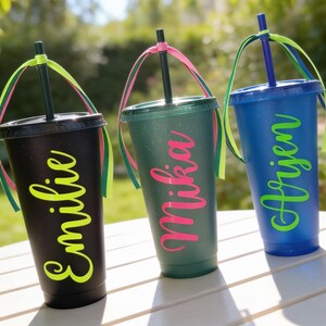 Puede incluir: Tres vasos de pl&aacute;stico reutilizables con tapa y pajita, cada uno con un nombre personalizado en letra cursiva. El vaso negro tiene el nombre en verde lima, el verde en rosa intenso y el azul en verde lima. Cada pajita est&aacute; decorada con cintas de colores.