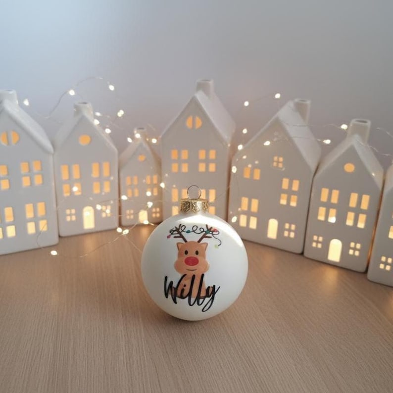 May include: A white Christmas bauble featuring a reindeer design and the name "Willy". It is set before a row of white ceramic house-shaped candle holders. A string of fairy lights is draped around the houses, creating a festive scene.