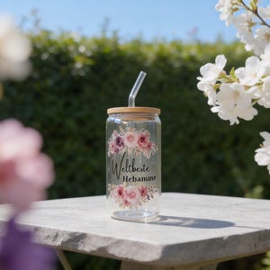 May include: A clear glass tumbler with a bamboo lid and a glass straw. The glass features a floral design in shades of pink and purple, with the black text "Weltbeste Hebamme" printed in the center between the floral arrangements.