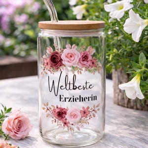 Op de afbeelding: Een doorzichtig glazen drinkglas met een bamboe deksel en een glazen rietje. Het glas is voorzien van een bloemmotief met roze en donkerrode rozen en de zwarte tekst "Weltbeste Erzieherin" in het midden, omringd door bloemstukken boven en onder.