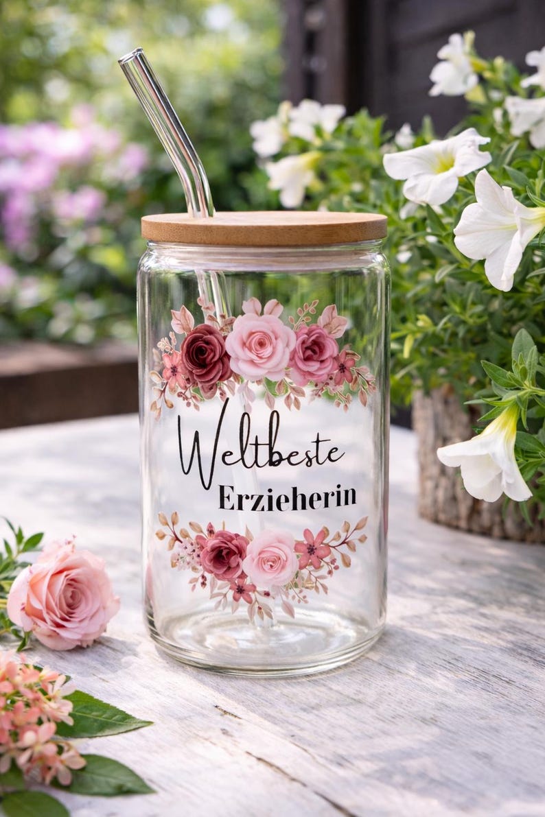 Op de afbeelding: Een doorzichtig glazen drinkglas met een bamboe deksel en een glazen rietje. Het glas is voorzien van een bloemmotief met roze en donkerrode rozen en de zwarte tekst "Weltbeste Erzieherin" in het midden, omringd door bloemstukken boven en onder.