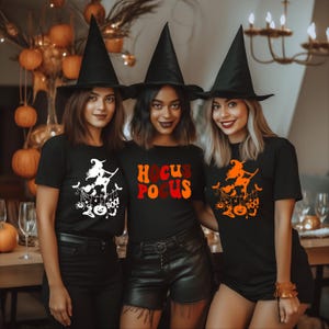 Könnte beinhalten: Drei Personen tragen schwarze T-Shirts und Hexenhüte. Die Shirts haben Halloween-Motive, darunter eine Hexe auf einem Besen, die Worte "Hocus Pocus" und einen Kürbis. Im Hintergrund sind Kürbisse und ein Kronleuchter zu sehen.