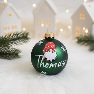 K&ouml;nnte beinhalten: Dunkelgr&uuml;ne Weihnachtskugel mit einer Zwerg-Illustration mit roter M&uuml;tze und wei&szlig;em Bart. Der Name "Thomas" ist in wei&szlig;er Schrift geschrieben. Wei&szlig;e Schneeflocken sind verstreut. Die Kugel hat eine goldene Kappe.