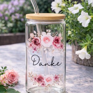 Pode incluir: Um copo transparente com tampa de bambu e palhinha de vidro. O exterior apresenta um design floral de rosas cor-de-rosa e brancas com folhas verdes, e a palavra "Danke" impressa em cursivo preto entre os arranjos florais.