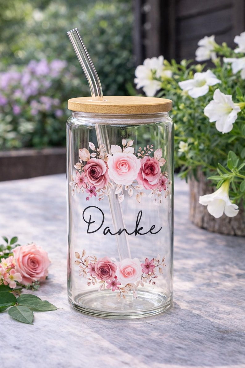 Pode incluir: Um copo transparente com tampa de bambu e palhinha de vidro. O exterior apresenta um design floral de rosas cor-de-rosa e brancas com folhas verdes, e a palavra "Danke" impressa em cursivo preto entre os arranjos florais.