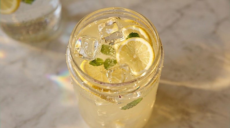 Pode incluir: Um copo transparente cheio de uma bebida c&iacute;trica amarelo-p&aacute;lida, com cubos de gelo, fatias finas de lim&atilde;o e folhas de hortel&atilde; verde fresca. A bebida &eacute; gaseificada com bolhas vis&iacute;veis e o copo tem uma borda lisa e arredondada.