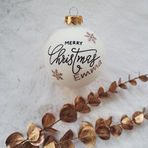 以下が含まれることがあります： 金色のアクセントと「Merry Christmas Emma」の文字が黒い筆記体で書かれた白いクリスマスオーナメント。 金色の雪の結晶のデザインも特徴です。 金色の装飾的な枝が前景にあります。