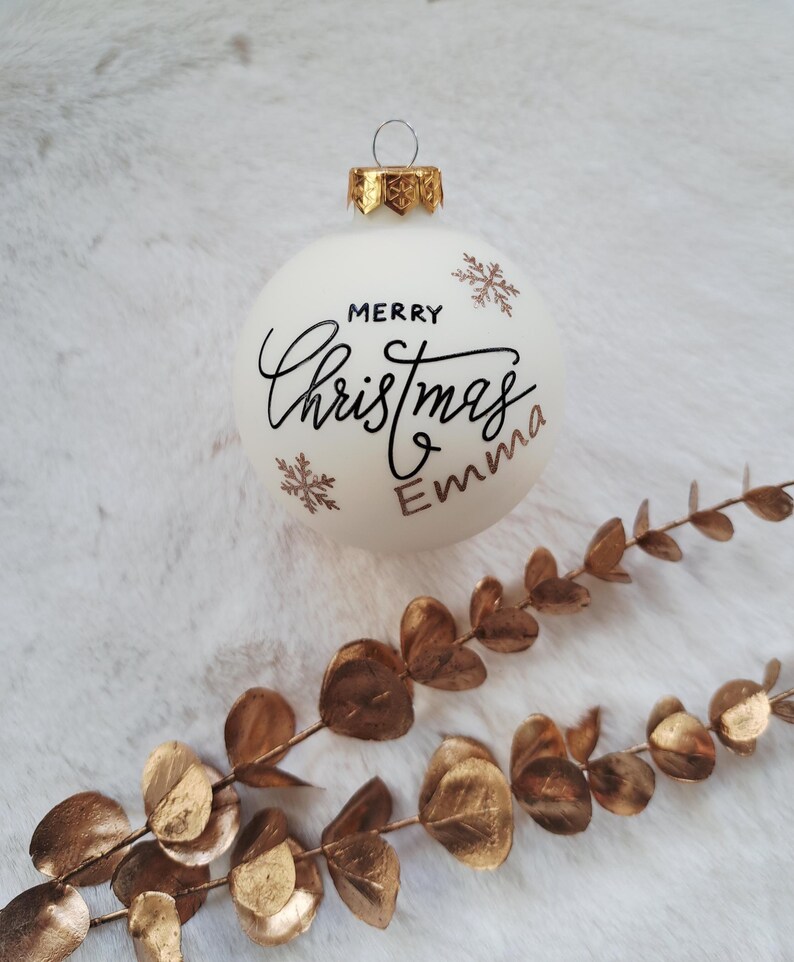 以下が含まれることがあります： 金色のアクセントと「Merry Christmas Emma」の文字が黒い筆記体で書かれた白いクリスマスオーナメント。 金色の雪の結晶のデザインも特徴です。 金色の装飾的な枝が前景にあります。