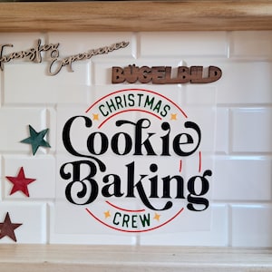 Könnte beinhalten: Ein gerahmtes Design zum Thema Weihnachten mit den Worten "Christmas Cookie Baking Crew" in einer kreisförmigen Grafik. Der Rahmen enthält auch Holzausschnitte mit der Aufschrift "Transfer Experience" und "Bügelbild" sowie sternförmige Verzierungen.