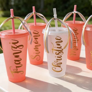 Puede incluir: Cinco vasos de pl&aacute;stico reutilizables con tapas y pajitas, cuatro en color coral y uno en blanco, cada uno con un acabado brillante y letras doradas en cursiva. Los nombres Franzi, Simone, Christine, Martina y Katharina est&aacute;n impresos.