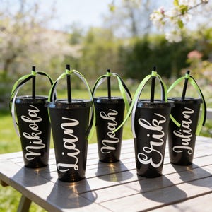 Puede incluir: Cinco vasos de pl&aacute;stico negro mate con tapas y pajitas negras, cada uno personalizado con un nombre en letra blanca: Nikola, Lian, Noah, Erik y Julian. Cada pajita est&aacute; decorada con una cinta de color verde lima y blanco atada arriba.
