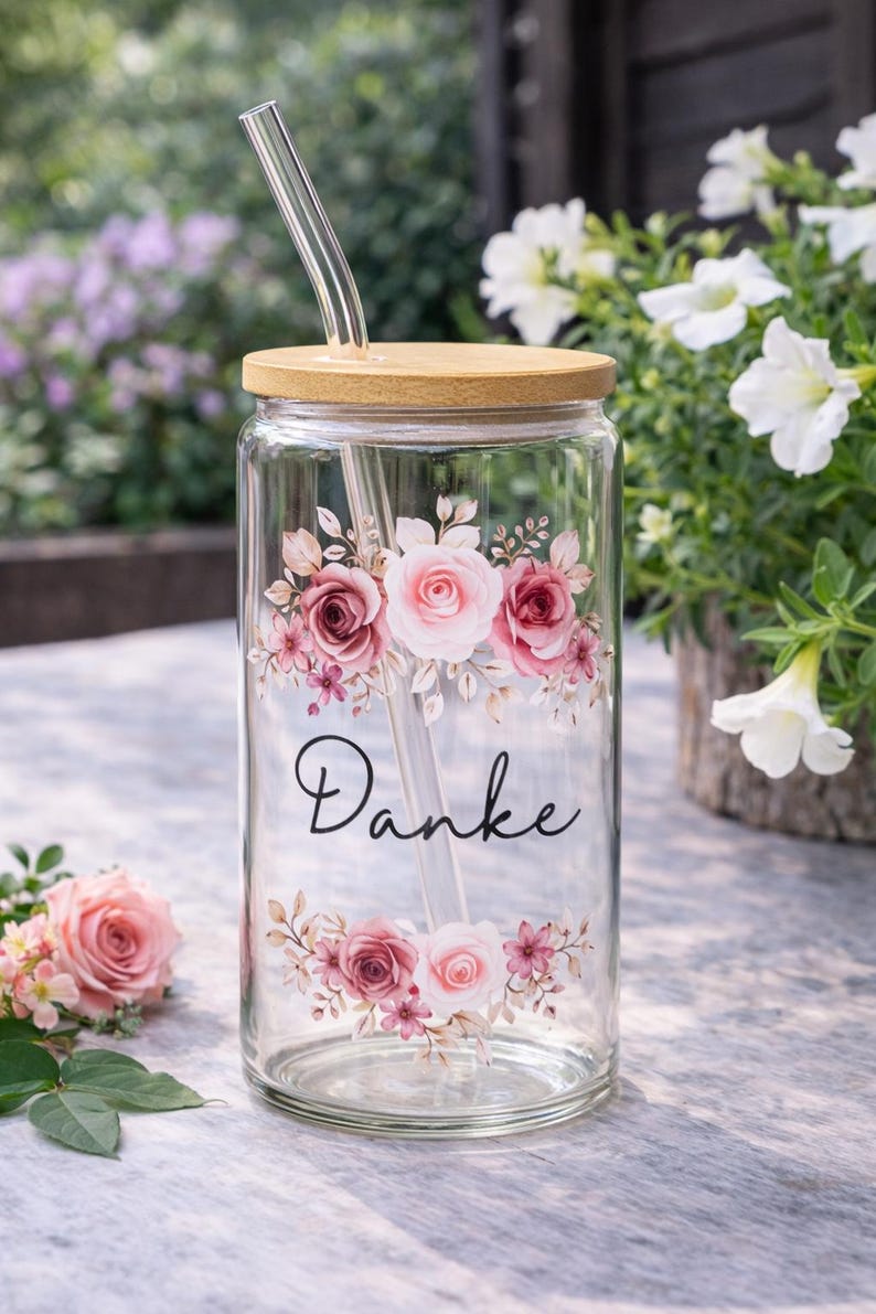 Op de afbeelding: Een doorzichtig glas met een bamboe deksel en een glazen rietje. De buitenkant is voorzien van een bloemmotief van roze en witte rozen met groene bladeren, en het woord "Danke" in zwarte cursieve letters tussen de bloemstukken gedrukt.