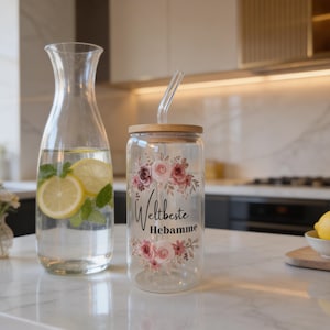 May include: A clear glass tumbler with a bamboo lid and a glass straw, featuring a floral design in shades of pink and the black text "Weltbeste Hebamme" printed on the front. The glass is positioned next to a clear glass water carafe.