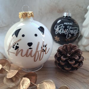 May include: White Christmas bauble with a dog face design and the name "Knuffi" in gold glitter. A black bauble with "Merry Christmas" in white script is also visible, along with a pinecone and gold leaf garland.