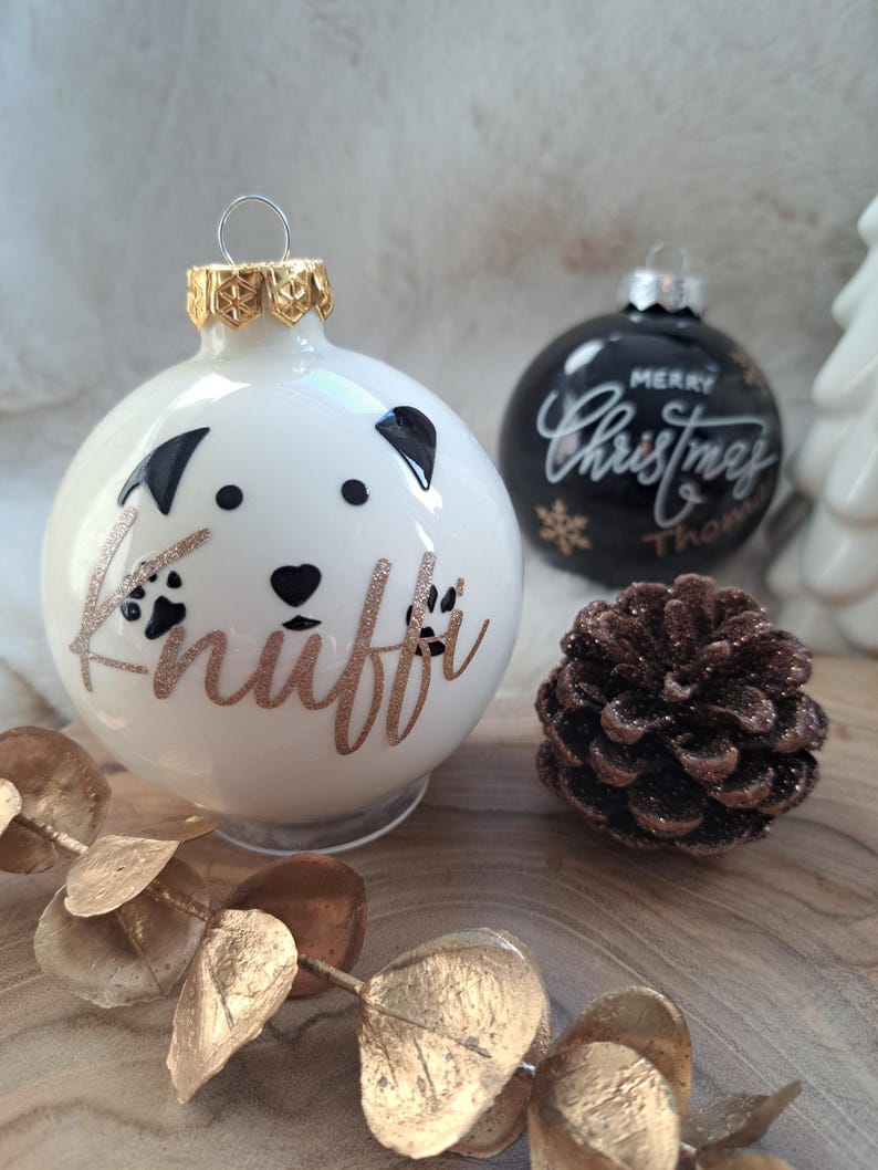 May include: White Christmas bauble with a dog face design and the name "Knuffi" in gold glitter. A black bauble with "Merry Christmas" in white script is also visible, along with a pinecone and gold leaf garland.