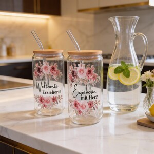 Pode incluir: Dois copos de vidro transparente com tampas de bambu e palhinhas de vidro, decorados com motivos florais cor-de-rosa. Um copo apresenta o texto "Weltbeste Erzieherin" e o outro "Erzieherin mit Herz" em letra preta. Um jarro de vidro e um lim&atilde;o est&atilde;o por perto.