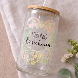 Pode incluir: Um copo de vidro transparente com tampa de bambu e canudo de vidro. O vidro apresenta um design de coroa floral em estilo aquarela em tons de rosa e verde, com o texto em alem&atilde;o "LIEBLINGS Erzieherin" impresso no centro da coroa.