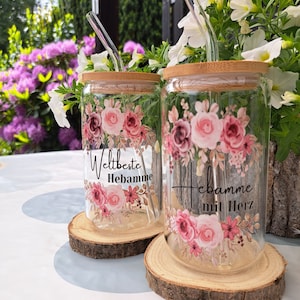 May include: Two clear glass tumblers with bamboo lids and glass straws, each decorated with pink floral patterns. One glass features the text 'Weltbeste Hebamme' and the other displays 'Hebamme mit Herz' in black script lettering.