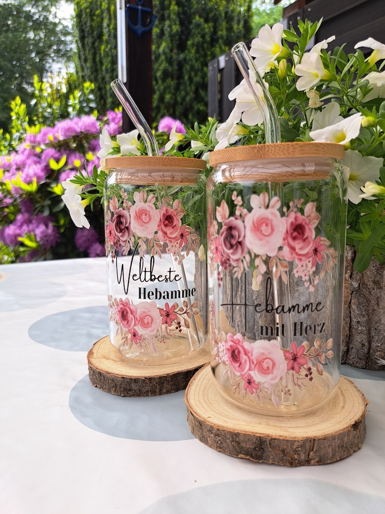 May include: Two clear glass tumblers with bamboo lids and glass straws, each decorated with pink floral patterns. One glass features the text 'Weltbeste Hebamme' and the other displays 'Hebamme mit Herz' in black script lettering.