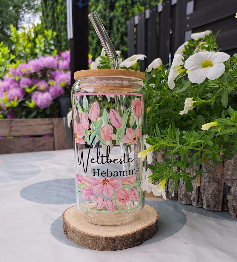 May include: A clear glass tumbler with a bamboo lid and a curved glass straw. The glass features a pink tulip floral pattern and the black text "Weltbeste Hebamme" printed in the center. The design is framed by thin pink and green lines.