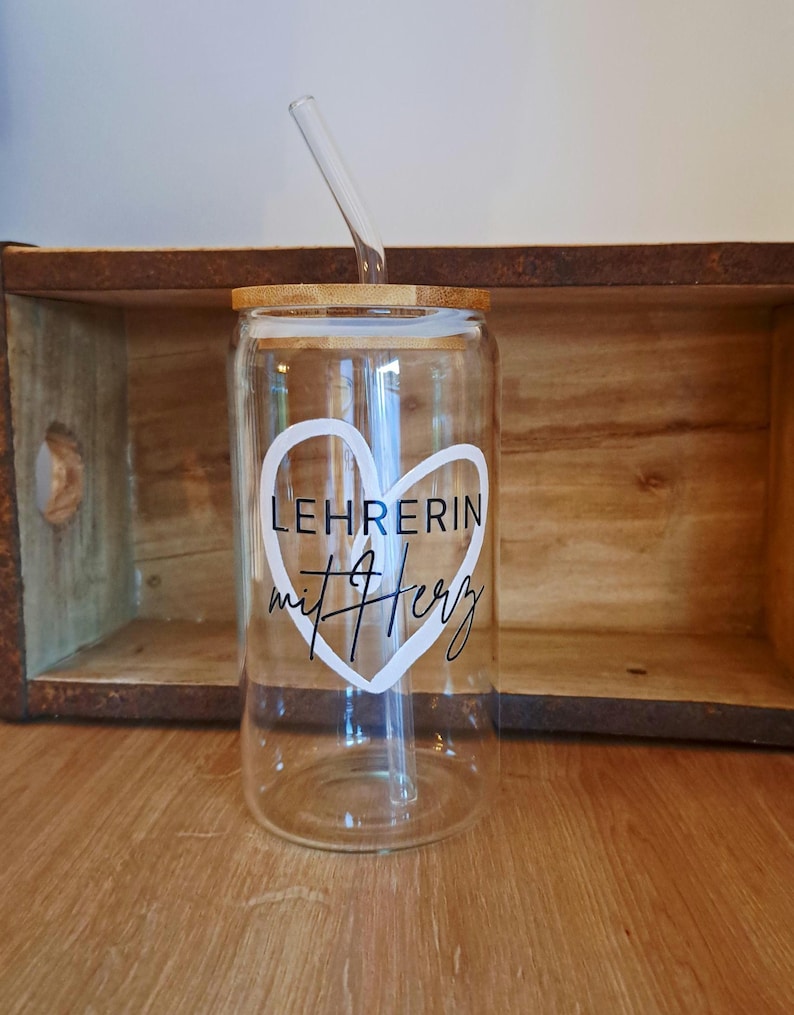 K&ouml;nnte beinhalten: Ein klares Glastrinkglas mit Bambusdeckel und Glasstrohhalm. Auf der Vorderseite befindet sich eine wei&szlig;e Herz-Grafik mit dem schwarzen Schriftzug LEHRERIN mit Herz im Inneren. Das Glas hat eine zylindrische Form mit einem flachen Boden.