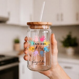 May include: A clear glass tumbler with a wooden lid and a straw. The tumbler features colorful handprints and the text "ERZIEHERIN mit Herz" and the name "Angelika".