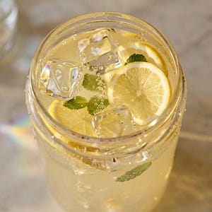 Op de afbeelding: Een helder glas gevuld met een lichtgele citrusdrank, met drijvende ijsblokjes, dunne schijfjes citroen en verse groene muntblaadjes. De drank is koolzuurhoudend met zichtbare belletjes en het glas heeft een gladde, ronde rand.