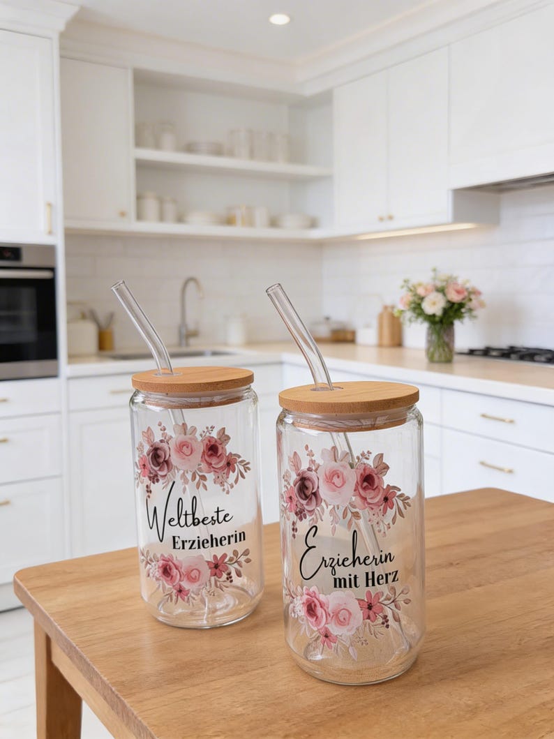 Op de afbeelding: Twee doorzichtige glazen bekers met bamboe deksels en glazen rietjes. Elke beker heeft een bloemmotief in roze en bruine tinten, met zwarte tekst "Weltbeste Erzieherin" op de ene en "Erzieherin mit Herz" op de andere beker.