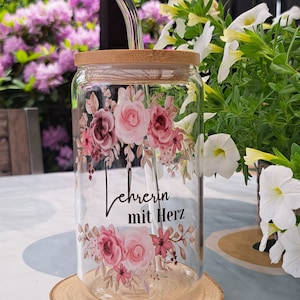 K&ouml;nnte beinhalten: Ein klares Glastrinkglas mit Bambusdeckel und gebogenem Glasstrohhalm. Das Glas ist mit einem Blumenmuster in Rosa- und Burgundt&ouml;nen verziert, wobei der schwarze Schriftzug "Lehrerin mit Herz" mittig zwischen den Blumenarrangements steht.