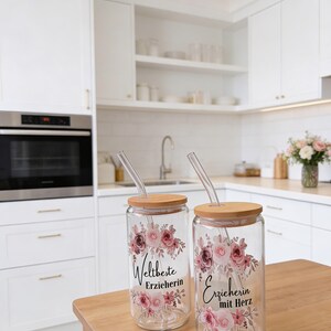 Pode incluir: Dois copos de vidro transparente com tampas de bambu e palhinhas de vidro, cada um com um design floral em tons de rosa e bordeaux. O copo da esquerda apresenta o texto 'Weltbeste Erzieherin' e o da direita 'Erzieherin mit Herz'.