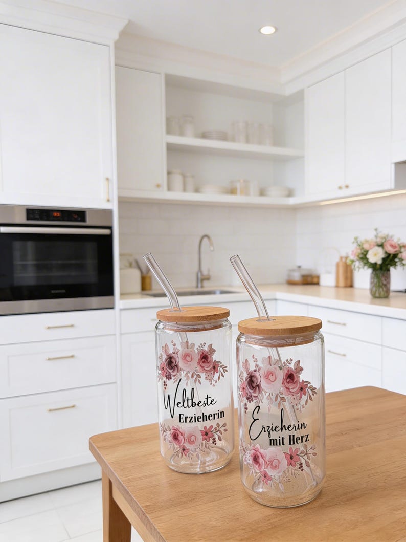 Pode incluir: Dois copos de vidro transparente com tampas de bambu e palhinhas de vidro, cada um com um design floral em tons de rosa e bordeaux. O copo da esquerda apresenta o texto 'Weltbeste Erzieherin' e o da direita 'Erzieherin mit Herz'.
