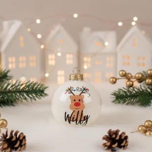 May include: A white Christmas bauble featuring a reindeer design and the name "Willy" in black script. The bauble has a gold-coloured metal cap and is surrounded by pine needles, gold berries, and miniature white houses. Festive holiday decor.