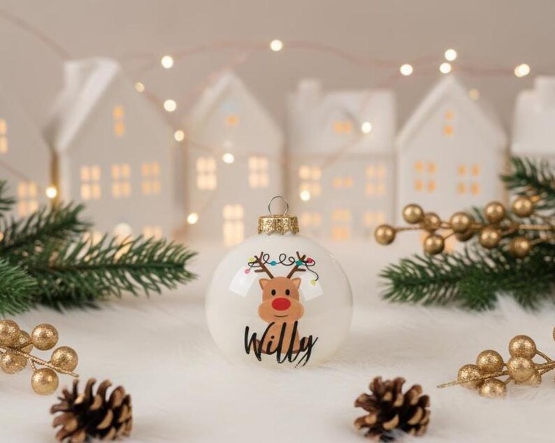 May include: A white Christmas bauble featuring a reindeer design and the name "Willy" in black script. The bauble has a gold-coloured metal cap and is surrounded by pine needles, gold berries, and miniature white houses. Festive holiday decor.